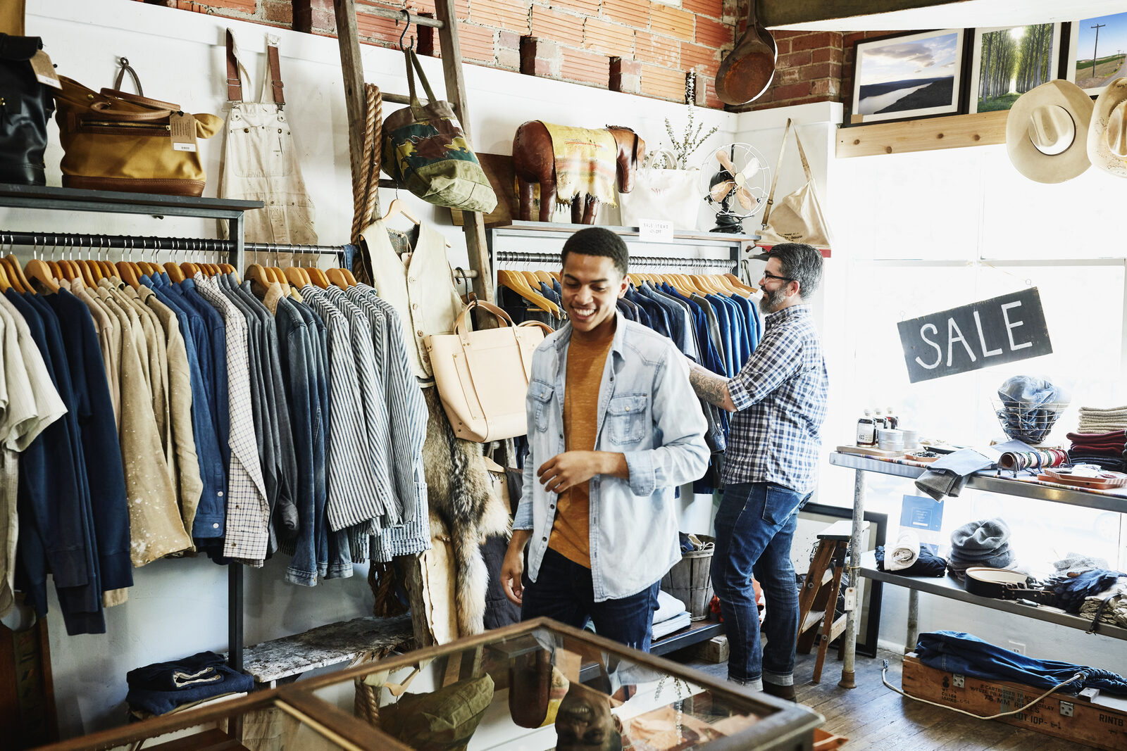 Two people in a clothing store, one browsing shirts, another smiling. SALE sign in window. Earthy tones dominate, creating a cozy vibe.