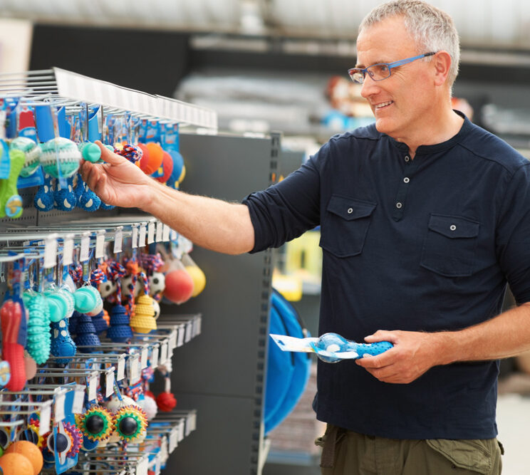 Man in a navy shirt shopping for colorful pet toys in a store, holding one toy and smiling. Shelves filled with various toys.