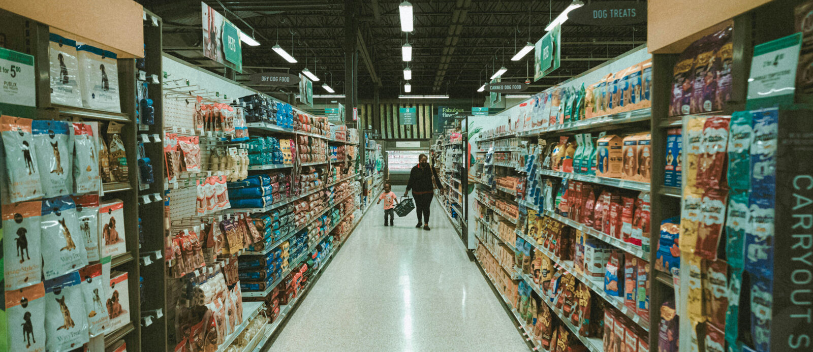 A person and child walk down a grocery aisle filled with pet food. Shelves display various colorful packages. Overhead signs read "Pets."