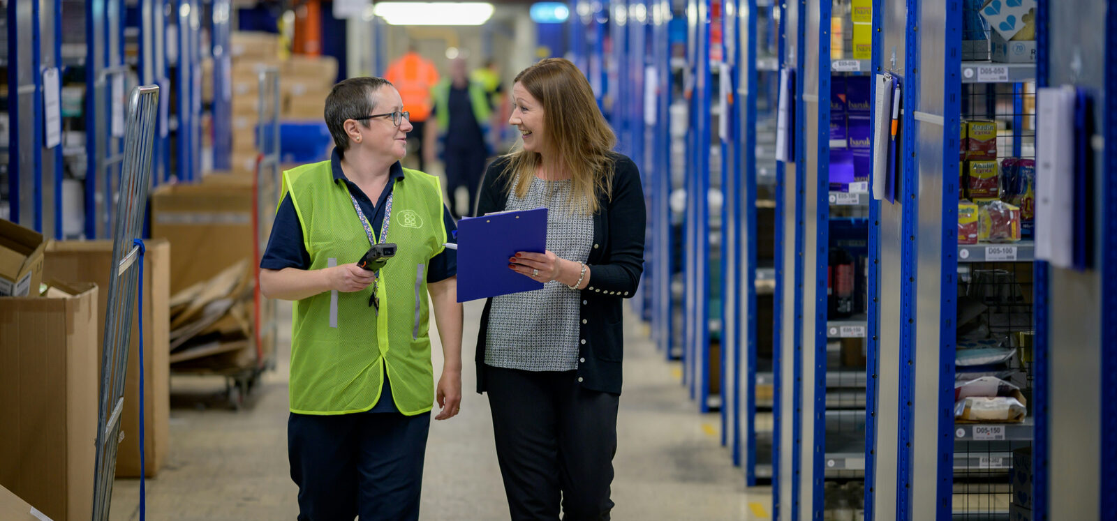 Two people in a warehouse walk and smile, one in a green vest holding a scanner, the other with a clipboard. Shelves with items line the aisle.